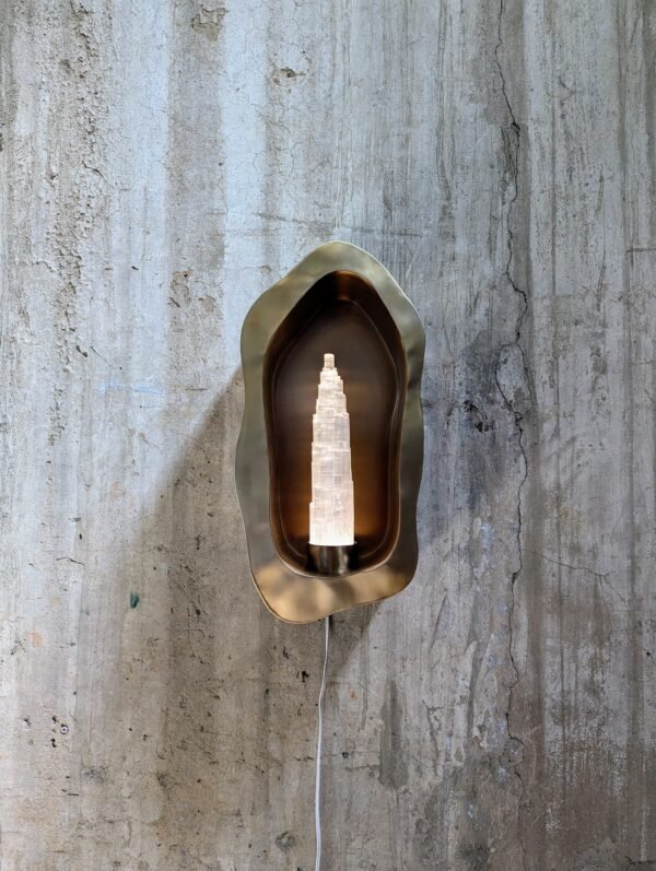 Front view of the Sigil Crest Wall Sconce showing the sleek stainless steel design and translucent selenite, mounted on a concrete wall.